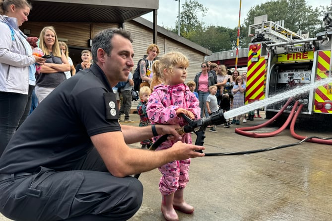 A youngster has a go at using a hose with a firefighter.  AQ 1977
